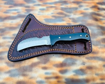 May include: A fixed-blade knife with a curved silver metal blade featuring a cross-hatched pattern and a smooth black handle with three brass pins. The knife rests on a dark brown leather sheath with decorative stamping and light stitching.
