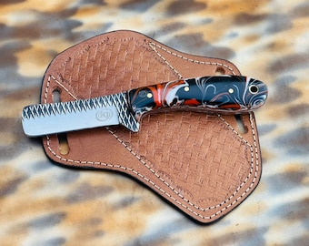 May include: A small, decorative knife with a patterned handle in black, red, and white rests on a brown leather sheath. The knife blade has a serrated edge and the sheath has a basketweave design with white stitching. The knife and sheath are displayed on a mottled background.