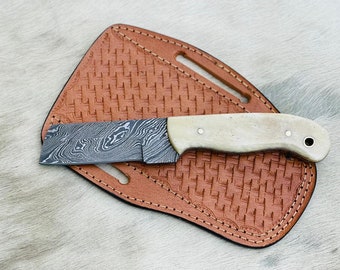 May include: A small, black and silver patterned knife with a white handle and a brown leather sheath.