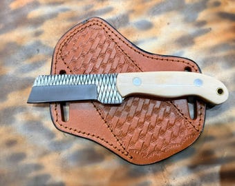 Puede incluir: Un pequeño cuchillo con una hoja plateada y un mango de madera clara descansa sobre una funda de cuero marrón. La funda tiene un patrón texturizado y está cosida en los bordes. El cuchillo tiene un agujero al final del mango.
