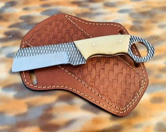 Puede incluir: Un cuchillo hecho a medida con un mango color crema y una hoja texturizada, sobre una funda de cuero marrón. La funda presenta un patrón tejido y bordes cosidos. El cuchillo tiene un anillo al final del mango.