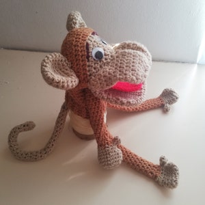 Puede incluir: Un títere de mono de crochet con pelo marrón y beige, ojos negros grandes y una boca roja abierta. El títere está sentado en un carrete de hilo.