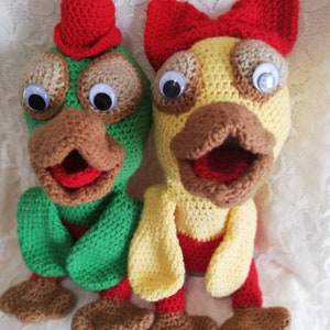 May include: Two crocheted stuffed toys shaped like ducks. One duck is green with a red hat and the other duck is yellow with a red bow. Both ducks have brown beaks and large white eyes.