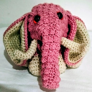 Può includere: Un giocattolo a forma di elefante all'uncinetto fatto a mano, rosa e beige. L'elefante presenta grandi orecchie flosce, una proboscide strutturata e occhi neri a bottone. Il giocattolo è realizzato con un punto stretto, che gli conferisce un aspetto robusto.
