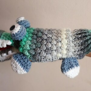 May include: Crocheted alligator puppet in shades of green, blue, and gray. The puppet features large googly eyes, a white-toothed mouth with a red tongue, and four blue and white feet. The body has a textured, bumpy pattern.