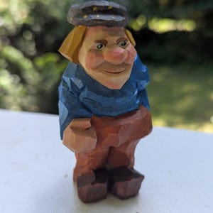 May include: A hand-carved wooden figurine of a smiling gnome wearing a blue shirt, brown pants, and a cap. The gnome has blonde hair and a rosy complexion. The figure is standing on a white surface with a blurred green background.