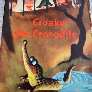 May include: A vintage children's book cover featuring a cartoon crocodile with blue eyes and a yellow and brown body. The title of the book is "Croaky the Crocodile" in red letters on a brown background.