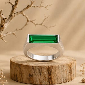 Peut inclure: Une bague argentée ornée d'une pierre précieuse rectangulaire vert émeraude, taillée en baguette et disposée horizontalement. L'anneau est lisse et poli, présentant un design géométrique épuré qui met en valeur la pierre verte éclatante.