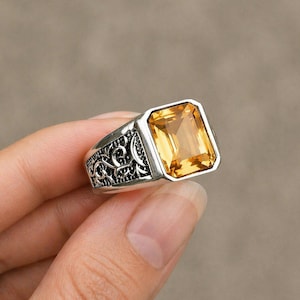May include: A silver ring with an ornate design on the band and a large, rectangular, yellow gemstone. The gemstone is set in a bezel setting, and the ring is held against a neutral background.
