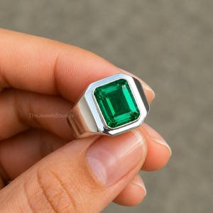 以下が含まれることがあります： 大きな長方形のエメラルドカットの緑色の宝石が付いたシルバーリング。リングは、洗練された仕上げのシンプルでモダンなデザインです。宝石はベゼルセッティングで設定されています。リングは手で持たれています。