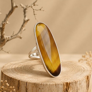 May include: A ring featuring a large, elongated oval tiger's eye gemstone set in a polished silver-toned metal band. The stone displays characteristic golden-brown chatoyant bands with a smooth, reflective surface and a tapered, vertical orientation.