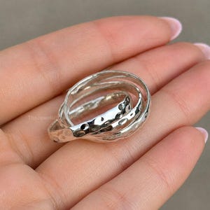 May include: A silver ring with three interlocking bands. The bands have a hammered texture, creating a unique, dimpled surface. The ring is displayed on a hand, showcasing its design and detail.