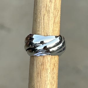 May include: A shiny, silver-colored ring with a woven design. The ring is displayed on a wooden dowel against a neutral background. The ring's surface reflects light, highlighting its intricate texture and shape.