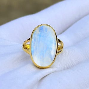 May include: A gold ring featuring an oval, iridescent moonstone. The moonstone has a pale blue and white color. The ring has a split band design. The ring is displayed on a white surface.