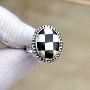Puede incluir: Un anillo de plata con un diseño ovalado a cuadros en blanco y negro. El anillo tiene un borde con cuentas y detalles intrincados en la banda. El patrón de tablero de ajedrez es un elemento de diseño clásico.