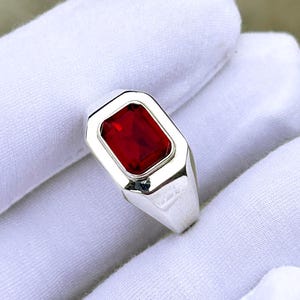 Puede incluir: Un anillo de plata con una piedra preciosa rectangular de color rojo intenso. El anillo tiene un diseño sencillo y moderno con un acabado pulido. La piedra preciosa está engastada en un bisel. El anillo se sostiene en una mano enguantada de blanco.