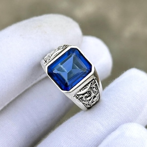 Op de afbeelding: Een zilveren ring met een grote, vierkante, diepblauwe edelsteen. De ring heeft sierlijke details op de band. De edelsteen is gezet in een vierkante bezel. De ring wordt vastgehouden door een gehandschoende hand.