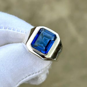 以下が含まれることがあります： 大きな長方形の濃い青色の宝石をあしらったシルバーリング。 宝石は、幾何学的なデザインの磨かれたシルバーバンドにセットされています。 リングは白い背景に表示されています。
