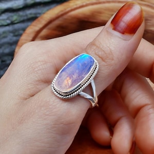 Puede incluir: Un anillo de plata con una gran piedra de luna ovalada engastada en un engaste de plata. El anillo tiene una banda delicada y retorcida.