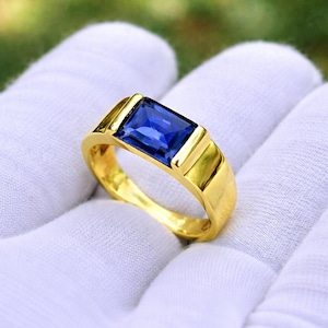 Peut inclure: Une bague en or avec une pierre précieuse rectangulaire bleu foncé. La bague a un design simple et élégant avec une finition polie. La pierre est sertie dans un sertissage à lunette, et le jonc est lisse et arrondi.