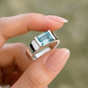 May include: A silver-toned ring featuring a rectangular, emerald-cut light blue gemstone set in a smooth, polished metal band. The stone is positioned horizontally within a tension-style setting, showcasing a clear, faceted surface and bright blue hue.