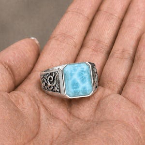 Può includere: Un anello d'argento con una pietra preziosa rettangolare azzurra. L'anello presenta intricati disegni in filigrana scura sui lati. La gemma è di colore azzurro chiaro con venature bianche. L'anello è mostrato su una mano.