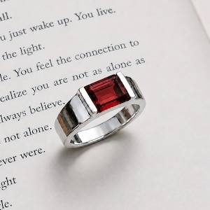 May include: A silver-toned ring featuring a rectangular, deep red gemstone set in a tension-style mounting. The band has a smooth, polished finish and is positioned on a page of a book with printed text visible in the background.
