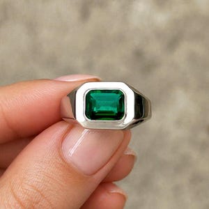 Könnte beinhalten: Ein silberner Ring mit einem rechteckigen, smaragdgrünen Edelstein. Der Ring hat ein klassisches Siegelring-Design mit poliertem Finish. Der Edelstein ist in einer Zargenfassung gefasst.