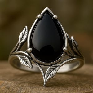May include: A silver ring featuring a large, teardrop-shaped black gemstone. The ring has a detailed, nature-inspired design with leaf accents on the band and small, round embellishments near the gemstone.