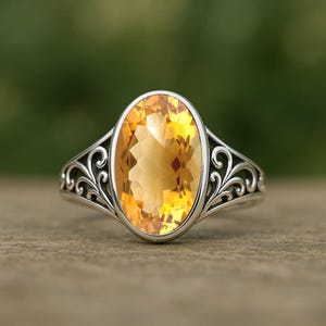 Puede incluir: Anillo de plata con una piedra preciosa ovalada de color amarillo dorado. La piedra preciosa está engastada en una banda de plata decorativa con un diseño de filigrana calada en los laterales. El anillo se muestra sobre una superficie de madera.