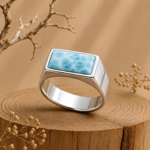 May include: A polished silver ring featuring a rectangular, light blue Larimar stone with a marbled, cloud-like pattern. The band is smooth and solid, showcasing a clean, metallic finish that contrasts with the textured, sky-blue gemstone inlay.