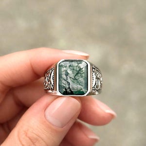 May include: A silver ring with a rectangular green stone. The stone has a unique, natural pattern. The ring has an ornate, detailed design on the band. The ring is held up by a person's hand.