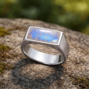 Könnte beinhalten: Ein silberner Ring mit einem rechteckigen, schillernden Edelstein in Blau- und Weißtönen. Der Ring liegt auf einem Stein, mit einem verschwommenen grünen Hintergrund. Schlichtes und elegantes Design.