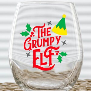 Puede incluir: Una copa de vino de vidrio transparente con un diseño rojo y verde que dice "The Grumpy Elf". El diseño también incluye un gorro de elfo verde con un borde amarillo y hojas de acebo.