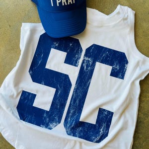 May include: A white tank top with large blue "SC" lettering and a blue trucker hat. The hat has white text that reads "YOU PLAY I PRAY". The tank top and hat are displayed on a neutral surface.