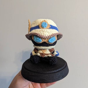 May include: A crocheted amigurumi figure of a character wearing a white and blue hat with a blue eye patch. The figure is sitting on a black circular base.