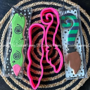 May include: Three cookie cutters in clear plastic packaging. The cutters are shaped like a witch's hat, a witch's broom, and a green witch's leg. The cutters are pink.