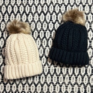 Może przedstawiać: Dwa dzianinowe zimowe czapki z pomponami z imitacji futra. Jedna czapka jest czarna, a druga kremowa.