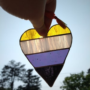 Peut inclure: Un cœur en vitrail aux couleurs du drapeau de la fierté non binaire : jaune, blanc, violet et noir. Le cœur est tenu contre un ciel clair, avec un petit pompon orange attaché. Le cœur est fait de morceaux de verre individuels.