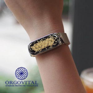 May include: A silver fitness tracker with a black and gold face featuring a geometric design. The text "ORGOVITAL ENERGIE PROTECTIE VITALITATE" is visible below the tracker.