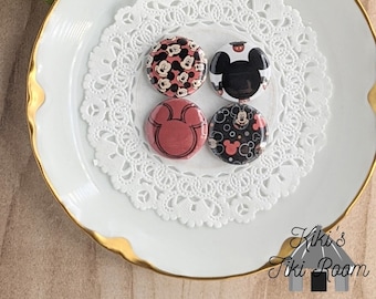 May include: Four round buttons with different Mickey Mouse designs. The buttons are on a white doily on a white plate with a gold rim. The text "Kiki's Tiki Room" is visible in the bottom right corner of the image.