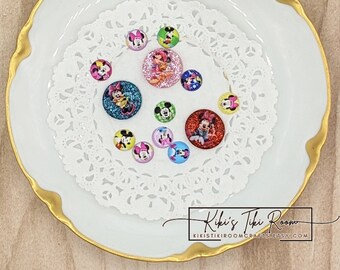 May include: A selection of colourful, round Disney-themed embellishments featuring Minnie and Mickey Mouse characters. The embellishments are arranged on a white plate with a gold rim and a decorative paper doily. The image includes the text "Kiki's Tiki Room".