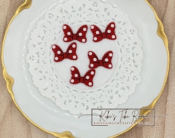 May include: Five red felt bows with white polka dots are arranged on a white lace doily, which is placed on a white plate with a gold rim. The image is from Kiki's Tiki Room.