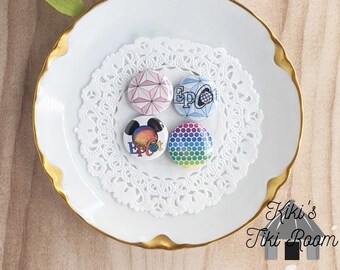 May include: Four round buttons with different designs. The buttons are on a white doily on a white plate with a gold rim. The buttons feature a rainbow design, a Mickey Mouse design with the words "Epcot", a blue and white design with the words "Epcot", and a pink and white geometric design.