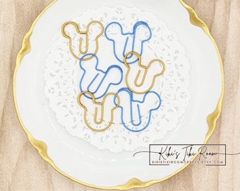 May include: A white plate with a gold rim holds a set of blue and gold metal paper clips shaped like Mickey Mouse ears. The paper clips are on a white doily. The text "Kiki's Tiki Room" and "kikistikitiroomcrafts.etsy.com" is printed on the plate.