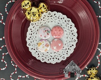 May include: Four round buttons with a white doily on a red plate. The buttons feature a pink and white snowflake design, a pink heart design, a pink and white Mickey Mouse design, and a colorful Christmas design with Mickey Mouse and Minnie Mouse.