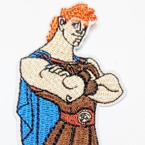 May include: Embroidered patch featuring a cartoon depiction of Hercules in a brown skirt, sandals, and a blue cape. The patch has a white border and the text "Kiki's Tiki Room Jewelry".