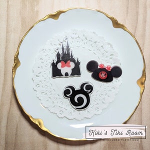 Peut inclure: Trois autocollants en silhouette de personnages Disney noirs et blancs sur un doily blanc. Les autocollants représentent Mickey Mouse, Minnie Mouse et le château de Cendrillon. Les autocollants sont sur une assiette blanche avec un bord doré.