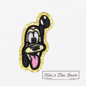 Op de afbeelding: Een strijk-op patch met een gouden glitter achtergrond met een cartoon hond met een roze tong die uitsteekt. De hond heeft zwarte ogen en een zwarte neus. De patch is ongeveer 5 cm hoog.