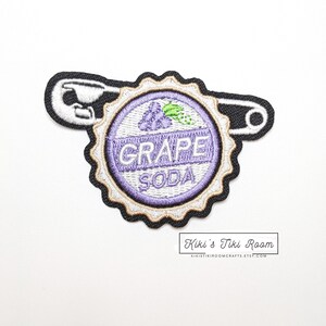 Könnte beinhalten: Gestickter Aufnäher in Form eines Grapefruitsoda-Flaschenverschlusses mit einer schwarzen Sicherheitsnadel. Der Aufnäher zeigt die Worte "GRAPE SODA" in weißer Schrift, eine lila Traube und ein grünes Blatt. Der Aufnäher hat einen weißen Hintergrund.